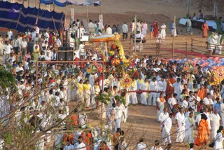 Photograph of an outdoor evening procession on a dusty ground with a decorated ratha-style palanquin at centre, its canopy and garlands in yellow, red and green. Hundreds of people in white dhotis and white saris walk around it, many with yellow scarves or saffron cloth, while drummers play drums at the lower right. Several people in orange and saffron robes walk among them. Striped and coloured parasols and a blue canopy stand overhead, with wooden barricades and tents behind. The palanquin's occupant is too small to identify.