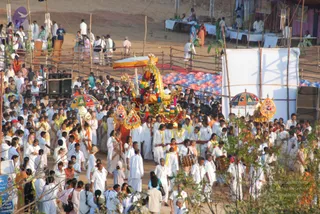 Outdoor festival procession on a dirt path lined with bamboo fencing and stalls. At the centre a decorated palanquin or ratha carries a deity murti under a yellow canopy, draped in garlands and colourful ornaments, with a figure in orange standing on it. Around it a dense crowd of devotees in white dhotis and white kurtas, many with yellow shawls, walks alongside drummers playing drums. Ornate umbrellas with multicoloured panels stand at the sides, and a long white screen and blue-covered stalls fill the background. The number of people is very large, but only the visible crowd is counted here.
