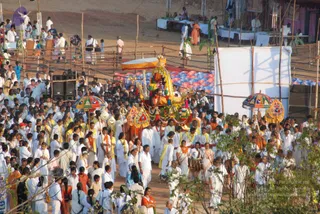 A large outdoor evening procession moves along a dirt path through a crowd of devotees dressed mostly in white, with some in orange and yellow. At the centre, a richly decorated ratha or palanquin carries a deity murti adorned with garlands, flower decorations and a golden crown-like canopy, with colourful umbrellas held over it. Drummers and attendants in white dhotis walk beside it. Bamboo fencing, tents, speakers and tables with white cloths line the open grounds, and a painted blue and purple pot display sits behind the procession. A figure in a bright orange robe stands at the upper right of the scene.