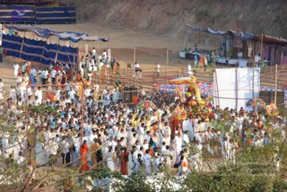 Outdoor evening procession on a dusty hillside site with blue tarpaulin awnings and bamboo fencing. A large crowd, mostly people dressed in white, walks around an ornate decorated palanquin or ratha carrying a deity murti, with a red-and-gold canopy and colourful garlands. Several people in white hold ceremonial staffs or umbrellas near the cart, and some devotees wear orange or saffron cloth. Stalls with tents and white banners line the background, and a tall rocky slope rises behind the scene.
