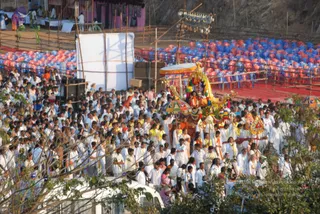 A large crowd of devotees, mostly dressed in white with some in yellow and orange, fills the foreground and follows an ornate decorated chariot (ratha) in a procession. The chariot has a gold and colourful flower-garlanded canopy with a yellow umbrella and a seated figure in orange and gold. Several white-clad attendants walk alongside holding decorative umbrellas and poles. Behind them, rows of red and blue plastic chairs fill a seating area beside a bamboo-framed stage and a white screen. Bamboo scaffolding and a rocky hillside form the background, with trees and a white van in the lower foreground.