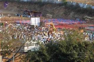 Elevated photograph of a large outdoor evening procession moving across a dusty ground past a hillside and white boundary wall. A dense crowd of people in predominantly white clothing fills the foreground, many with white garments and some in orange or pink. At the centre a decorated ratha-style float with a golden canopy and colourful fringed decorations is carried through the crowd. Rows of red and blue seated chairs or mats fill a fenced area on the right, with a white screen and a small canopied stall beside them. A white ambulance-style van with a red cross-like emblem stands at lower left, and strings of bunting hang on bamboo poles.