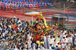 A large outdoor religious procession moves through a crowd of many people dressed in white with yellow sashes and garlands. At the centre, a decorated golden ratha or chariot carries a seated figure in orange-saffron robes with long dark hair, under a white canopy with gold finials and a multicoloured parasol nearby. The chariot is draped with yellow and red garlands and flower strings. Scaffolded seating with blue and red chairs and red mats frames the scene, and an orange flag with a blue emblem flies on the left. A standing figure in orange clothing walks beside the chariot.