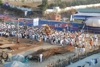 Aerial view of a large outdoor procession at an ashram-style grounds with a stage and blue-red mat area. A dense crowd of people dressed mostly in white moves behind an ornate colourful chariot or palanquin decorated with red, yellow and orange motifs and an umbrella-like canopy. Some women wear saris in pink, purple and orange. A striped tent and a stage with lights sit at right, and bundled bamboo poles and sand piles lie in the foreground. Many people are in white dhotis and shirts, and a few in saffron-orange robes walk near the chariot.