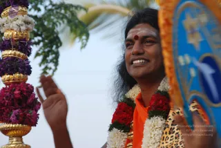 A smiling man with long dark hair and white and red tilak on his forehead stands outdoors beside a tall golden and flower-decorated ceremonial pole with purple and pink blossoms. He wears an orange saffron garment and garlands of red roses and white jasmine, and raises one hand while holding a blue and orange ceremonial fan-shaped object with a leopard-print cloth. A palm tree and pale sky are in the background. Swamiji (probable): the long hair, saffron clothing and floral garlands are the visible cues, and he is shown in person, celebrating during an outdoor procession.