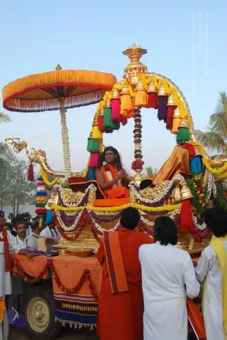 Outdoor procession on a golden ceremonial chariot decorated with marigold and jasmine garlands, colourful tassels and gold bells beneath a canopy, with a golden horse-head prow and a large yellow fringed parasol. A man with long dark hair, wearing an orange robe and rudraksha-style beads, sits centrally on the gilded seat with his hands folded in greeting. Beside him, a figure in orange drapes reclines on cushions. In the foreground, two people in white garments and one in a yellow shawl walk alongside the chariot, while a crowd of onlookers in white and orange stands at left near a decorated orange-draped cart. Palm trees and a clear sky form the background.