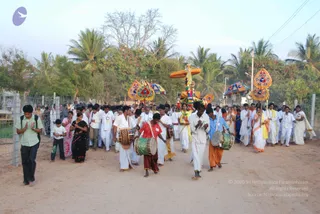 A village procession moves along a dirt road lined with palm trees and a wire fence. Several drummers lead, two playing large barrel drums with woven straps; one wears a red dhoti and another a blue shirt with orange dhoti. Behind them people in white dhotis and shirts and women in saris carry a decorated ratha or palanquin topped with an orange canopy and a kalasha-like finial, flanked by colourful ornate umbrellas. A young man in a green shirt and black trousers walks at the left with hands in anjali. Many devotees follow in white and saffron clothing.