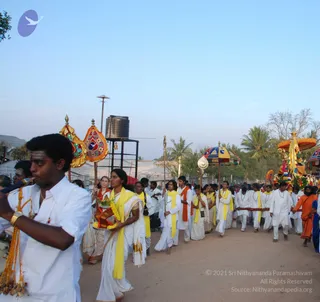 Outdoor evening procession on a dirt road with palm trees and a water tank beside a fence. Men and women in white clothes walk in a line; a woman in a white sari with a yellow shawl carries a brass tray with flowers and offerings. Several men carry tall staffs and decorated umbrellas and banners with gold and red embroidery. On the right, a decorated chariot or palanquin with yellow canopy and garlands carries a deity murti. A man in white with a rudraksha mala walks beside it, and a bearded man in orange robes stands at the right edge. A man at left plays a wind instrument with a gold-and-yellow cord. Several people wear yellow or orange shawls.