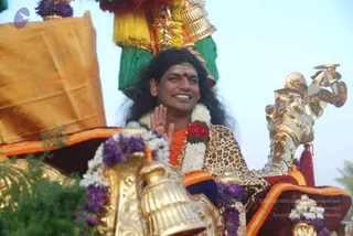 A man with long dark swept-back hair, an orange-saffron garment with a leopard-print shawl, and a white and red flower garland sits on an ornate golden procession vehicle with a gilded carved mount and a tall green, yellow and orange tassel canopy above. He smiles and raises a hand in greeting, with a red bindi and white tilak on his forehead. Purple and white flower garlands decorate the golden carriage, and a yellow-orange cloth drapes at left. Swamiji (probable): the visible cues are long swept-back hair, saffron clothing, flower garlands and a ceremonial procession setting, so this is the person on the chariot in procession.