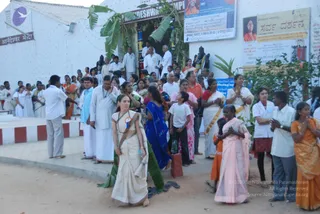 Outdoor evening scene in front of a white temple building with a banana-leaf doorway decoration and an entrance sign reading Nandeshwar temple. A woman in a cream sari with a green-and-gold border walks in the foreground, holding a water bottle and wearing a blue lanyard badge. Behind her, a long line of devotees in white dhotis and kurtas, saris in orange, pink, red and blue, and white ritual garments waits and stands with hands folded in namaste. A dark stone deity murti stands on the right near the temple wall, and a large banner on the wall shows a portrait and Kannada text reading Sarva Darshan. A bearded man in a white shirt stands at the left edge of the crowd.