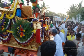 Outdoor evening procession on a palm-lined road. A richly decorated golden ratha/chariot covered in marigold, jasmine and multicolour flower garlands, with green foliage, bells and red tassels, carries a smiling man with long dark hair wearing an orange garment, a white shawl and flower garlands; he raises a hand in greeting. Around the chariot, many devotees in white clothes and yellow shawls walk and reach toward it, some holding decorated fans or flags. Several onlookers stand along the roadside and the crowd pushes forward. Swamiji (probable) is shown in the procession, seated on the chariot with long swept-back hair, orange clothing and garlands.