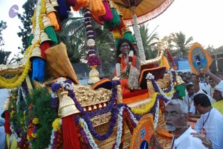 A richly decorated golden ratha procession chariot with a canopy of orange, yellow and green tassels and flower garlands of marigold and purple blooms. Seated on a red and gold cushioned throne at the centre is a smiling figure with long dark hair, wearing an orange robe, a leopard-print shawl, white floral garlands and a red bindi, raising one hand in blessing. Probable Swamiji by appearance: long hair, saffron robe and garlands, seated enthroned on the ratha. Around the chariot, men in white kurtas and dhotis walk, one in front with glasses and grey hair holding an orange and blue fan-shaped ceremonial ornament. Palm trees and a crowd stand in the background, with a banner reading Ananda Satsang nearby.