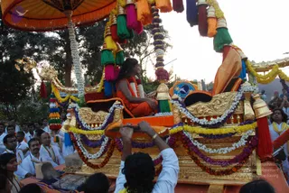A photograph of an evening procession with a richly decorated golden palanquin-style chariot draped in marigold, white and purple garlands, with colourful tassels hanging from a canopy above. A man with long dark hair, wearing an orange-saffron robe and a red-and-white garland, sits on the decorated seat, and a figure in a brown-orange shawl stands at the rear of the chariot. Around the chariot, devotees in white dhotis and white shirts walk beside it, some with yellow and gold sashes, and a man in white holds up an object with raised arms in the foreground. a person is seated in the chariot, identified by long swept hair, saffron robe and garlands, as its central occupant.