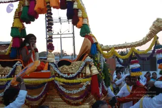 A photograph of an evening procession with a richly decorated golden ratha or chariot draped in marigold and jasmine garlands and tasselled orange, green, pink and red fabric canopies. A woman with long dark hair, wearing an orange garment and white flower garlands, sits on the chariot seat smiling and gesturing with her hand. Behind her, a man wearing an orange garment and a multicoloured tasselled headdress stands near the chariot, while people in white clothes and orange shawls walk alongside. A golden elephant-trunk shaped ornament extends at the right edge. a person candidate not established; the visible figure's appearance is uncertain.