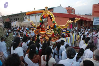 A photograph of a festival procession on a street beside an orange-walled building. A golden ratha-style chariot with a dragon-headed prow, a yellow canopy and hanging tassels, dangling bells and bright multicoloured fabric streamers is pulled through the crowd. A seated figure in saffron and orange robes rides atop the chariot under the canopy. Below, a dense crowd of devotees in white clothes and white dhotis, plus women in saffron and white saris, follows and presses around the vehicle, with some wearing yellow shawls. A signboard at right reads Nithyananda Charitable Dispensary. Multiple people, mostly in white, fill the foreground and midground.