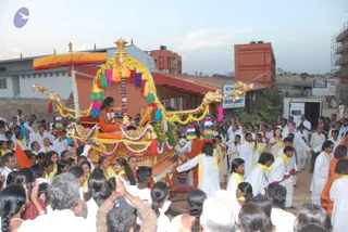 Outdoor evening procession on a street with a decorated golden palanquin carried through a crowd. A seated figure in orange robes with long dark hair sits under a yellow fringed canopy on the ornate golden vehicle, which is hung with garlands, multicoloured tassels and pink, yellow and purple streamers. Around it, many people in white clothes with yellow shawls walk and reach toward the palanquin, some with hands raised in greeting or folded in namaste. Devotees in the foreground have hands joined in prayer. Behind them are orange-roofed buildings, a sign reading Nithyananda Dhyanapeetha Charitable Dispensary, and a white van. Many people are visible across the scene.