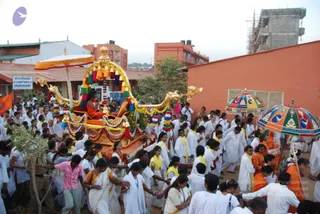 A festival procession moves through a dusty street past orange-brick buildings. At centre, a richly decorated golden palanquin with carved dragon-head ends, yellow and orange tassels and colourful garlands carries a seated figure in orange robes with a crown-like headdress, lifting a hand in blessing. Around it, a large crowd of devotees in white clothes with yellow shawls and sashes walks alongside, many holding hands in a chain. Two decorative parasols with blue, orange and green panels and gold finials shade the procession on the right, and a saffron flag is visible at left. A sign reads Nithyanada Charitable Dispensary. Under construction scaffolding and a partly built structure rise behind the scene.