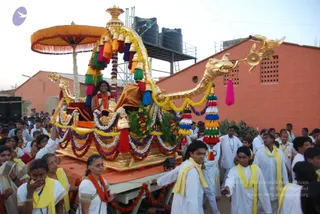 A photograph of an outdoor procession with a gilded golden ratha (chariot) carrying a decorated palanquin-style seat, topped by a yellow and orange canopy with fringe and a golden makara-headed prow. The chariot is draped with garlands of marigolds, white flowers and purple beads, and hung with multicoloured tassels and bells. A young person in orange-and-red clothing sits on the seat. Around the chariot, many people in white dhotis and kurtas with yellow shawls pull and walk alongside, while women in saris and yellow shawls follow. Red-brick buildings, water tanks and a scaffold stand behind, and a sound system is visible at left.