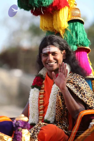 A smiling man with long dark hair and white and red tilak marks on his forehead sits in a procession vehicle, waving with his right hand. He wears an orange saffron robe, a leopard-print shawl, and white jasmine garlands with red flowers. A gold bead necklace hangs on his chest. Behind him rise a tall canopy with bright yellow, green, red and pink tassels and a gold finial. Blurred purple flowers and a gold lamp edge the lower left. Swamiji (probable): long swept-back hair, saffron robe, rudraksha-style gold beads and a ceremonial garland mark the figure as a probable Swamiji in a procession.