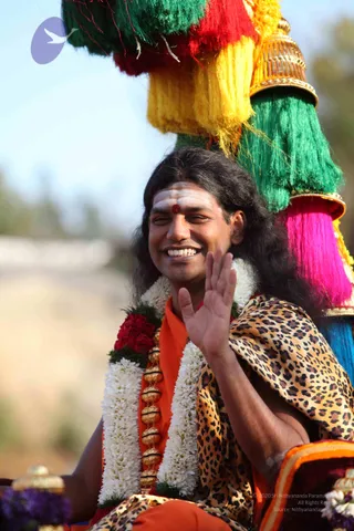 A smiling man with long dark hair sits on a decorated procession vehicle, waving his right hand. He wears an orange saffron garment, a leopard-print shawl over one shoulder, white and red flower garlands, and a gold-beaded necklace. His forehead bears white vibhuti lines and a red kumkum dot. Behind him rise tall tassels in green, red, yellow, pink and blue, mounted on a gold-capped pole, against a bright blue sky.
