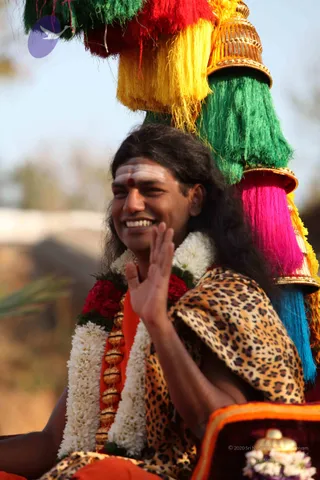 A smiling man with long dark hair, white sacred ash on his forehead and a red dot at the centre, sits on a procession vehicle waving his right hand. He wears a leopard-print shawl over an orange garment and a double strand of white jasmine garlands with red flowers, plus a gold-beaded rudraksha-style mala. Behind him rises a tall ceremonial canopy topped with a gold finial and hung with bright tassels in red, yellow, green, magenta and blue. Dry grass and a pale sky fill the background, and a red velvet cushion with flowers sits at the lower right. a person appears as this figure, identified by long swept hair, ornate gold-beaded mala, orange clothing and garlands, seated on the ceremonial procession vehicle.