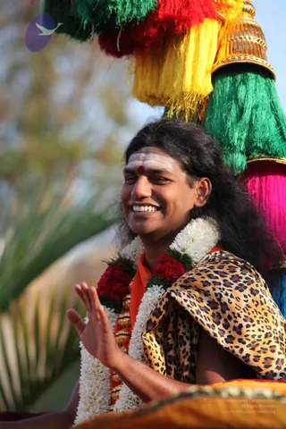 A smiling man with long dark hair, white vertical tilak marks and a red bindi on his forehead, sits under a colourful canopy of red, yellow, green and magenta tassels topped with a gold finial. He wears white and marigold-style garlands of white flowers and red roses, an orange garment, and a leopard-print shawl over his shoulders, and raises one hand in greeting. The background is blurred foliage in daylight. Swamiji (probable) appearance cues: long swept-back hair, ornate garlands and saffron-orange clothing, seated in a procession setting.