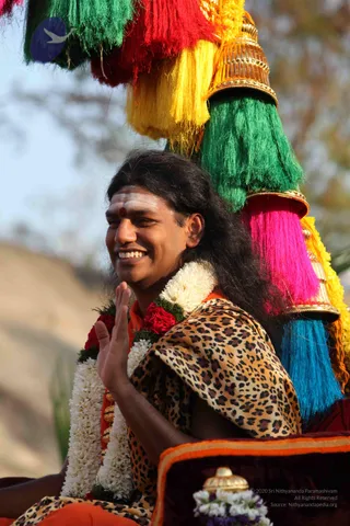 A smiling man with long, swept-back dark hair sits on an ornate palanquin or ratha beneath tall fringed tassels in green, red, yellow, pink and blue topped with gold finials. He wears a leopard-print shawl over orange clothing, a white and red flower garland (jasmine and roses) around his neck, and a white forehead marking with a red dot. He raises one hand in greeting. Background is blurred trees and sky.