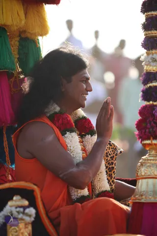 A man with long dark hair sits on a decorated ratha or palanquin, smiling and holding his palms together in anjali mudra. He wears an orange sleeveless top and a white and red flower garland over his shoulders, with a leopard-print cushion behind him. Colourful tassels in yellow, green, pink and teal hang on the left, and a gold-and-flower decorated pillar stands on the right. Blurred figures in light clothing stand in the background under bright sunlight.
