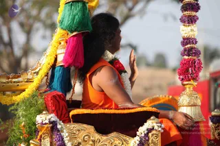 A photograph of a person seated in a decorated golden palanquin or ratha during a procession, viewed from behind and slightly to the side. The person wears an orange sleeveless top and has long dark hair, with a white and red floral garland draped on the shoulders. The raised right hand is open in a blessing gesture toward the unseen crowd. The golden carved seat has a maroon velvet cushion with orange trim. The canopy above is decorated with yellow marigold garlands, green, pink and teal tassels, and a vertical pole wrapped in purple, pink and white flowers. Outdoors, with dry trees and a red vehicle in the background.