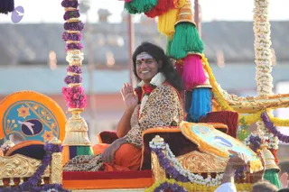 A photograph of a procession float decorated with purple and white flower garlands, gold ornaments and colourful tassels in green, pink, blue and yellow. A man with long dark hair and a red forehead mark sits on a gold-framed seat wearing a leopard-print garment over orange saffron cloth and a white flower garland, smiling and raising one hand in greeting. Hands of devotees reach toward the float at the bottom edge. Swamiji (probable): the seated man has long swept-back hair, saffron clothing and a ceremonial garland, consistent with the page's reference to Swamiji at this evening procession. Visible people include the seated man and partially visible hands in the crowd.