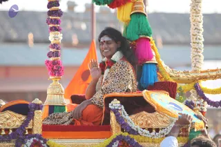 A man with long swept-back dark hair sits in a richly decorated golden palanquin during a procession, waving his right hand and smiling. He wears a leopard-print shawl over orange clothing, a rudraksha-style red and white garland, and a white tripundra mark with a red dot on his forehead. The palanquin is draped with purple, white and yellow flower garlands and has bright green, pink, blue and red tassels hanging from its canopy. An orange flag stands behind him, and a hand holds a decorated fan at the lower right. Swamiji (probable): the long hair, leopard shawl, orange clothing and garlands match the presentation, seated in the procession's palanquin.