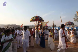 An outdoor evening procession on a dusty ground under a pale sky, with hills and unfinished buildings behind. Many devotees in white clothes and white dhotis walk in a crowd. In the centre, a decorated palanquin-style ratha is shaded by a multicoloured umbrella with tassels, its carved golden figures and ornaments visible. Two men carry long staffs topped with gold finials, and a second colourful umbrella stands at right. A woman in a blue sari walks beside the palanquin. A man at right in white holds a staff with an orange cloth. Blue portable toilet units line the background. Visible people number roughly twenty to thirty.