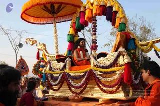 A photograph of a golden ceremonial palanquin-style chariot (ratha) during a procession, decorated with yellow, purple, white and red garlands and a large orange canopy with fringed tassels. A man with long dark hair, wearing an orange robe and a white flower garland, sits cross-legged on the golden seat in a meditative pose. Below, a shallow basket holds fruit and an orange cloth drapes the platform. Onlookers stand along the roadside, including a man on the left and a man in orange clothing at the right edge, with a woman in a red top partially visible in front. Swamiji (probable) is the seated figure, identified by long swept-back hair, orange robes and garland cues; he is seated in a golden chariot as the procession's central figure.