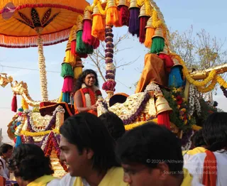 Outdoor festival procession with a golden ratha chariot decorated with marigold and jasmine garlands, bells, and tassels in red, pink, purple, green and yellow. A man with long dark hair, orange-saffron robe, white flower garland and red floral garland sits on the chariot seat, smiling. Orange canopy with fringe overhead. In the foreground, several people in white and yellow shirts with orange scarves walk and pull near the chariot. Probable Swamiji is seated on the decorated seat, identified by long swept-back hair, saffron robe and garlands.