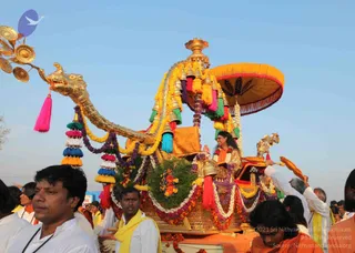 A decorated ceremonial chariot procession with a golden makara-headed prow and a large saffron canopy umbrella. A figure with long dark hair in a saffron and gold robe sits on the chariot, one hand raised in greeting, surrounded by garlands of marigolds and purple flowers and hanging tassels in pink, teal and yellow. Men in white kurtas with yellow shawls walk beside it, one in front with a rudraksha-style mala and vibhuti on his forehead, and a devotee in orange clothing stands at the lower right. Attendants on the chariot wave golden fans and a bell-topped staff. The procession moves under a clear blue sky.
