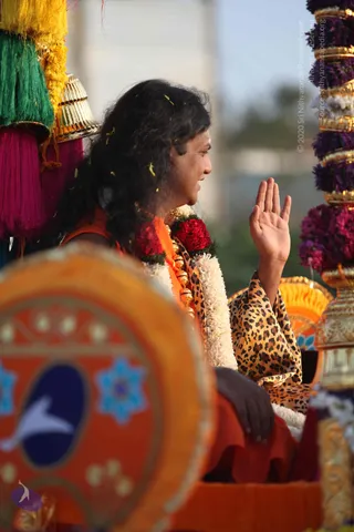 Photograph of a long-haired figure seated on an ornate chariot during a procession, raising one hand in greeting. The figure wears a leopard-print shawl over orange-saffron garments, adorned with white jasmine garlands and red flower garlands, and gold beaded necklaces. Behind and beside the figure stand tall pillars wrapped in purple and pink flowers and gold bands, with colourful green, yellow and magenta tassels hanging at left. In the foreground, an orange-red painted panel with a blue oval and star motifs is blurred. Swamiji (probable), seated on the chariot in a procession; the appearance cues are long swept-back hair, saffron clothing, rudraksha-style gold beads and garlands.