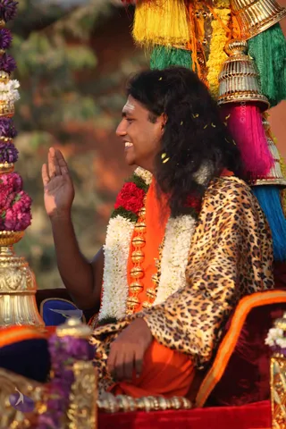 A photograph of a long-haired man seated on a richly decorated golden palanquin or chariot, waving his right hand to the side. He wears a leopard-print shawl over an orange-saffron garment, with white and red flower garlands and a gold rudraksha-style bead necklace. Behind him, colourful silk tassels in yellow, green, pink and blue hang from gilded finials, and purple and pink flower garlands decorate a gold pillar at left. Petals fall in the air during a procession.