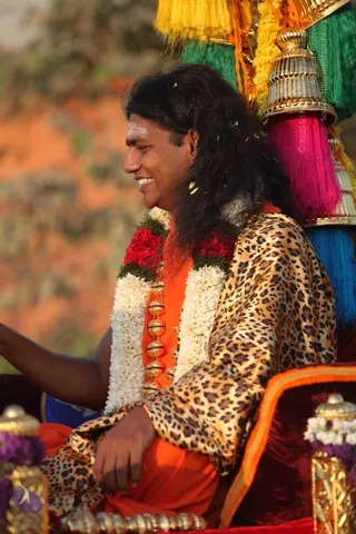 A smiling man with long, dark, wavy hair and a white-and-red tilak on his forehead sits on a richly upholstered seat during an outdoor procession. He wears an orange garment under a leopard-print satin shawl, and a necklace of white jasmine and red flower garlands hangs over his chest, with gold bead ornaments among them. Behind him rise a tall gilded finial and tassels in yellow, green, pink and blue, and loose petals scatter through the air. His left arm extends toward the left edge of the frame. Orange-brown rocky terrain fills the blurred background.