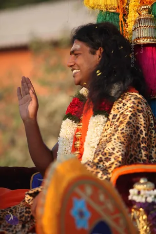 A smiling man with long, swept-back dark hair rides in a procession, raising his right hand in greeting. He wears a leopard-print shawl over an orange-saffron garment and a garland of white jasmine and red flowers around his neck, with a gold necklace visible. A white tilak mark is on his forehead. Behind him stands a decorated golden and pink tasselled ornament on a palanquin-like structure, with yellow petals scattered in the air. A blurred orange and blue cushion or decorative cloth fills the lower foreground. Swamiji (probable): long swept-back hair, saffron garment and gold necklace on a procession vehicle, with a greeting gesture.