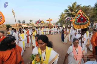 A photograph of an outdoor religious procession on a dirt road lined with palm trees, with a low sun and hazy sky behind. Devotees in white dhotis and white saris with yellow and pink shawls walk in a long line. Ceremonial parasols with red, gold and floral embroidered canopies are carried aloft, and a decorated palanquin or ratha with garlands is visible in the middle distance. In the foreground, a bearded man in orange robes walks at left, and a woman in a white sari with a yellow shawl carries a brass kalasha-style vessel wrapped with flowers and leaves. A woman in white with glasses and a pink shawl walks at right. Ceremonial staffs and a golden trident-like standard are held by marchers, and some people carry umbrellas. The overall count of visible people is large.