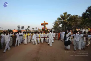 A dirt road procession moves forward at dusk under a pale sky with palm trees on the right. A decorated ratha or palanquin, topped with an orange canopy and floral garlands, is carried by men in white dhotis and white shirts with yellow shawls and red sashes. Crowds of devotees in white saris and white shirts walk alongside, some with yellow scarves, and a few figures in orange robes appear near the right. Several men in white shirts and trousers hold their arms outstretched to guide the crowd. Palm trees and a building with red walls stand in the background.