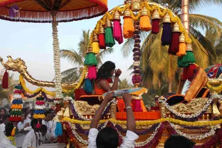 A photograph of an ornately decorated golden palanquin procession under palm trees. On the raised golden seat, a long-haired figure wearing an orange-saffron garment sits with hands raised in greeting or blessing. The palanquin is draped with garlands of white and purple flowers, hanging tassels in green, red, orange, yellow and magenta, and a gold-carved elephant-headed finial at the front. Above, an ornate umbrella with fringe shades the seat. Below, a man in a light shirt raises both hands toward the seat, and several white-clad devotees and bystanders stand around the chariot in the crowd.
