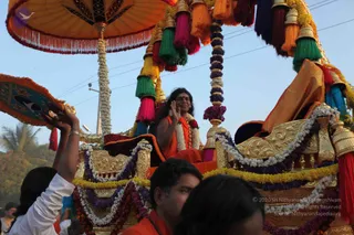A photograph of an evening procession. A man with long swept-back dark hair, wearing an orange saffron garment and a white flower garland with a rudraksha-style mala, sits on an ornate gold chariot and raises one hand in greeting. The ratha is decorated with garlands of white and purple flowers, hanging tassels in pink, green, orange and blue, and a large orange canopy overhead. Devotees in white and orange clothing stand around the chariot, one raising an arm near a decorated fan or parasol. Palm trees and power lines appear against a pale evening sky.