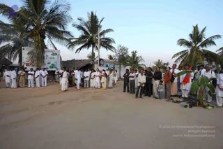 Outdoor photo of a sandy ground lined with tall coconut palms at dusk, with a large information board on the left reading in part Nithyananda and a thatched-roof structure behind. A long line of people stands facing the same direction: many men and women in white ritual clothes and white shirts, some women in saffron and orange saris, and men in dark shirts and light trousers on the right. A small child stands near the right group. Footwear is piled on the ground near a banana plant at right, and a red and white flag is visible. Statues stand near the right edge beside the crowd.