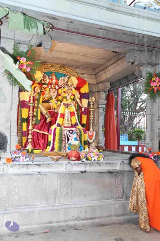 A photograph of a temple shrine with a deity murti pair seated on a raised platform, adorned with marigold, yellow and red flower garlands and pink gerbera blossoms. A brass shivalinga-like murti sits in front with garlands and a small blue-clad figure. Brass oil lamps stand on either side, with a brass plate of offerings and a red vessel on the stone platform. In the lower right, a person in an orange robe and a leopard-print shawl bows deeply, head lowered toward the shrine. Behind the shrine, saffron cloth hangs in an open pillared hall with trees beyond. A banana-leaf decoration hangs at the top left, and the photo is watermarked with copyright text.