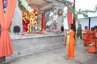 A photograph of an outdoor temple shrine with a grey stone platform and pillars draped in orange cloth and banana leaves. A large deity murti is decorated with flower garlands in red, yellow and purple, and a brass lamp stands in front. A long-haired figure in orange robes with a leopard-print shawl stands with palms joined in prayer facing the shrine. To the right, several people in orange robes and rudraksha malas hold brass plates and offerings, and a brass tray with a lit flame sits on a stand.
