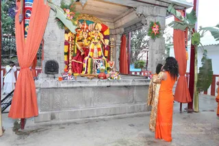 A photograph of a temple shrine under an open pavilion with orange cloth drapes and banana leaves. Inside, a deity murti pair is decorated with yellow and orange marigold garlands and a flower-garlanded backdrop, with brass lamps and flower offerings in front. A woman in an orange sari stands with hands raised in prayer at the shrine, beside a person in a patterned leopard-print wrap. A photographer in white stands at left by a tripod. Three people are visible.