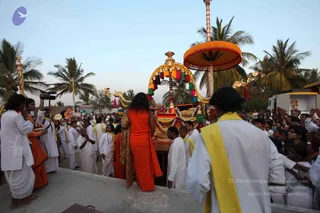 Outdoor evening procession under palm trees. A long-haired figure in a saffron-orange robe with a leopard-print shawl walks with their back to the camera, beside an ornate golden palanquin with a red canopy, green-and-gold tassels and gilded serpent-like finials. Devotees in white dhotis and white kurtas, several wearing yellow shoulder cloths, walk and stand around it, some with hands raised, and a crowd watches from the right. An orange parasol-style umbrella rises above the palanquin, and a small shrine-like structure with a deity figure appears at the far right.