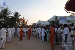 Outdoor evening procession on a dirt road lined with palm trees. A decorated chariot (ratha) with an orange canopy and garlands moves at the left, while a crowd in white ritual clothes, many with yellow shawls, walks alongside. Two saffron-robed figures walk in the foreground: one woman in orange sari and one man in orange robe, with the orange-robed man holding a shawl. Devotees in white kurtas and dhotis form a line holding hands near the right. A whitewashed building with hanging banners and a printed portrait stands behind the crowd, and a red and gold umbrella with tassels frames the right edge.