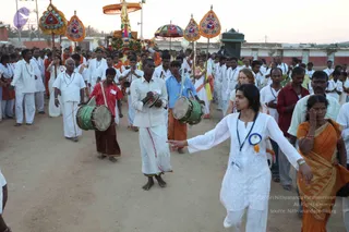 A photograph of an evening procession on a dirt road with a decorated ratha or palanquin-style canopy at the top of the frame, topped by a golden umbrella-like structure. Ornate triangular teardrop-shaped parasols in orange, red and gold with blue and white accents are carried on poles. A large crowd of over a hundred people in white dhotis and kurtas with rudraksha-style necklaces walks along. In the foreground a woman in white churidar with a blue rosette badge points forward with an outstretched arm. Two drummers in white and red clothes play large green-and-brown drums, and a man in a white shirt plays cymbals. A woman in an orange saree wipes her face at the right edge. Walls and palms are visible in the background at dusk.